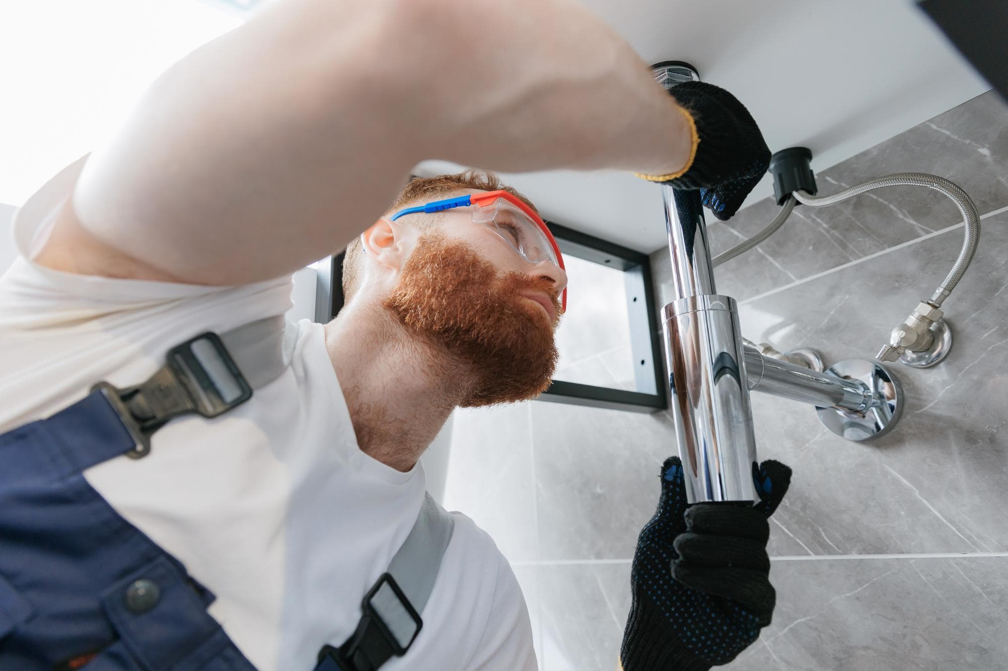 A plumber wearing protective goggles and gloves is fixing a shiny metal pipe under a sink in a modern bathroom. Captured from a low angle, the photo highlights his focused expression and pipe repair Paramus plumbing tools.