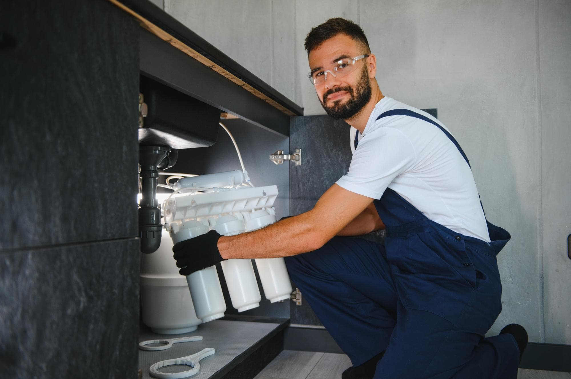 A plumber in a white shirt, blue overalls, gloves, and safety glasses installs or repairs a water filtration system under a kitchen sink, smiling at the camera—showcasing professional Plumbing Services.