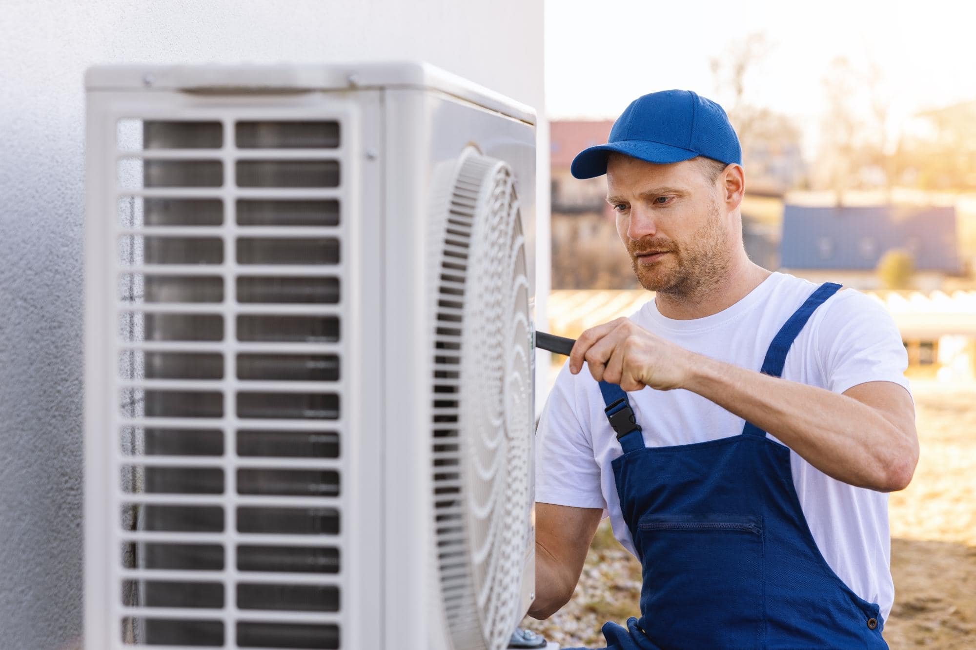 A technician in blue overalls and a blue cap uses a screwdriver to repair or maintain an outdoor air conditioning unit attached to a building, similar to the expertise offered by a plumbing company Hackensack residents trust.