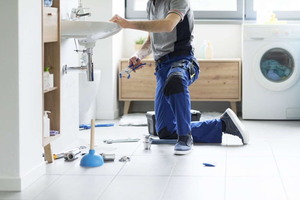 A plumber in blue work pants kneels on a bathroom floor, fixing pipes under a sink, with tools and a plunger nearby. A washing machine and wooden cabinet are in the background—perfect for pipe repair Paramus professionals.
