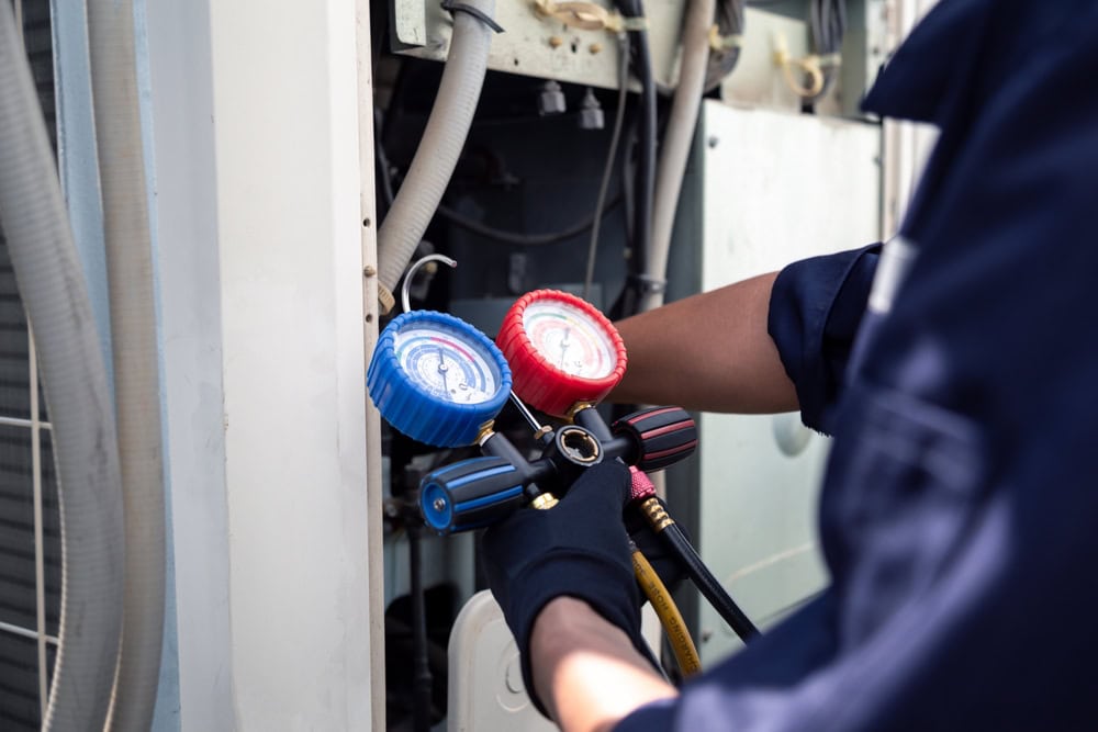 A technician wearing gloves adjusts blue and red gauges on HVAC equipment, checking or repairing an air conditioning unit as part of comprehensive Plumbing Services in Teaneck.
