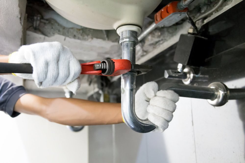 A person wearing white gloves uses a red pipe wrench to tighten a metal pipe under a sink, demonstrating expert plumbing services.