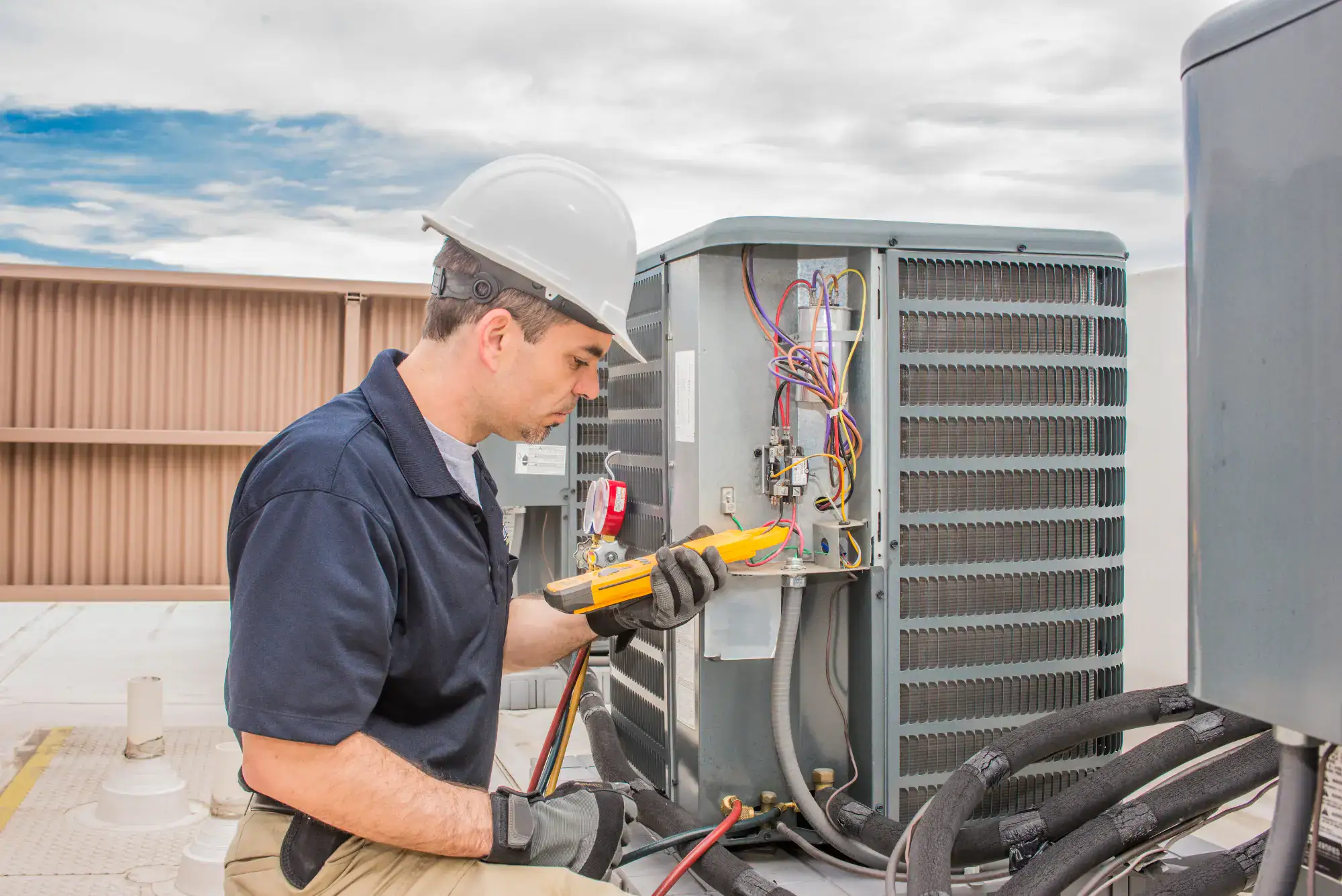 A technician wearing a hard hat and gloves uses a multimeter to inspect and repair wiring on an outdoor air conditioning unit on a rooftop.