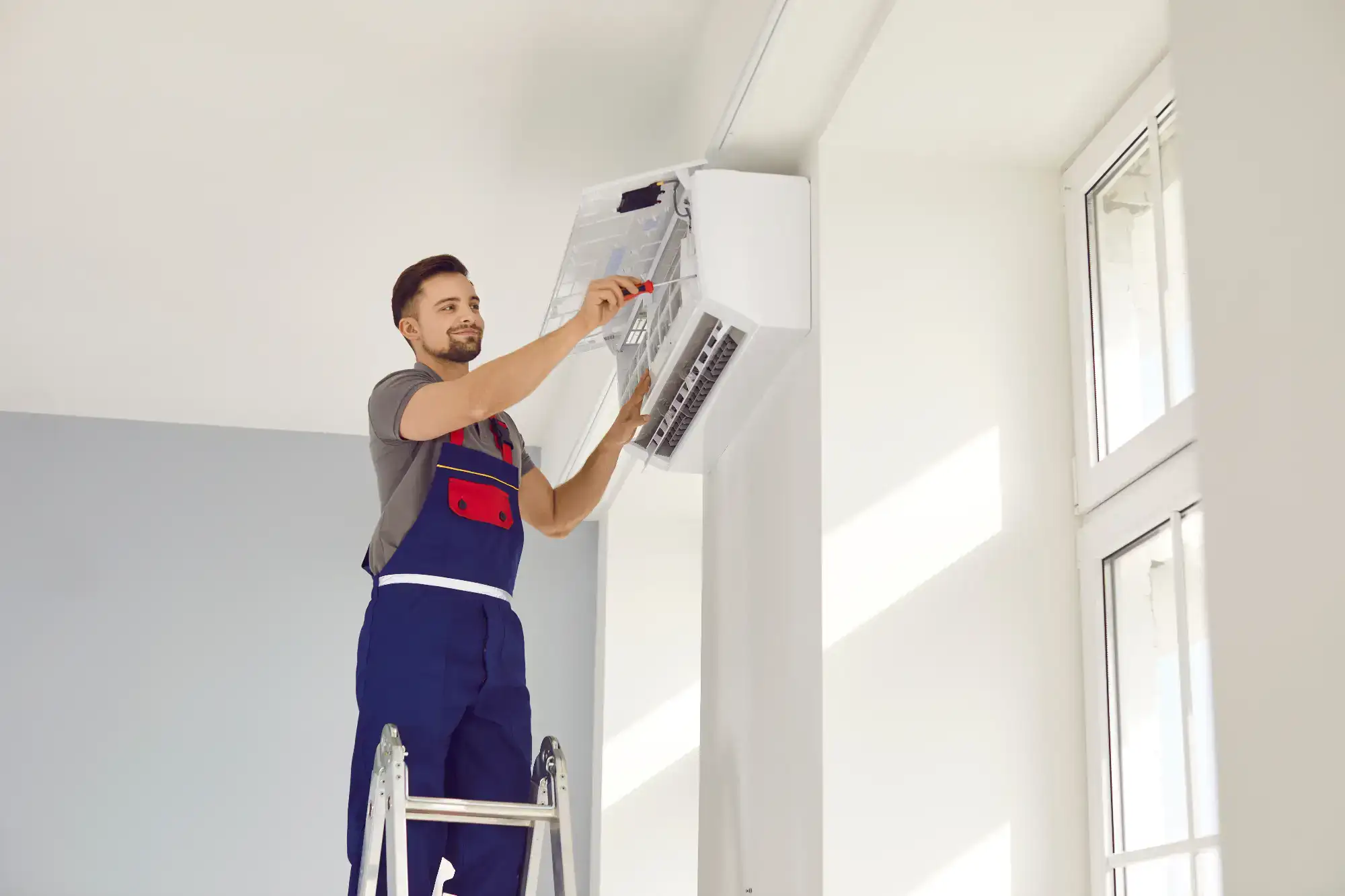 A technician in blue overalls stands on a ladder, repairing or installing a wall-mounted air conditioner unit in a bright, modern room with large windows.