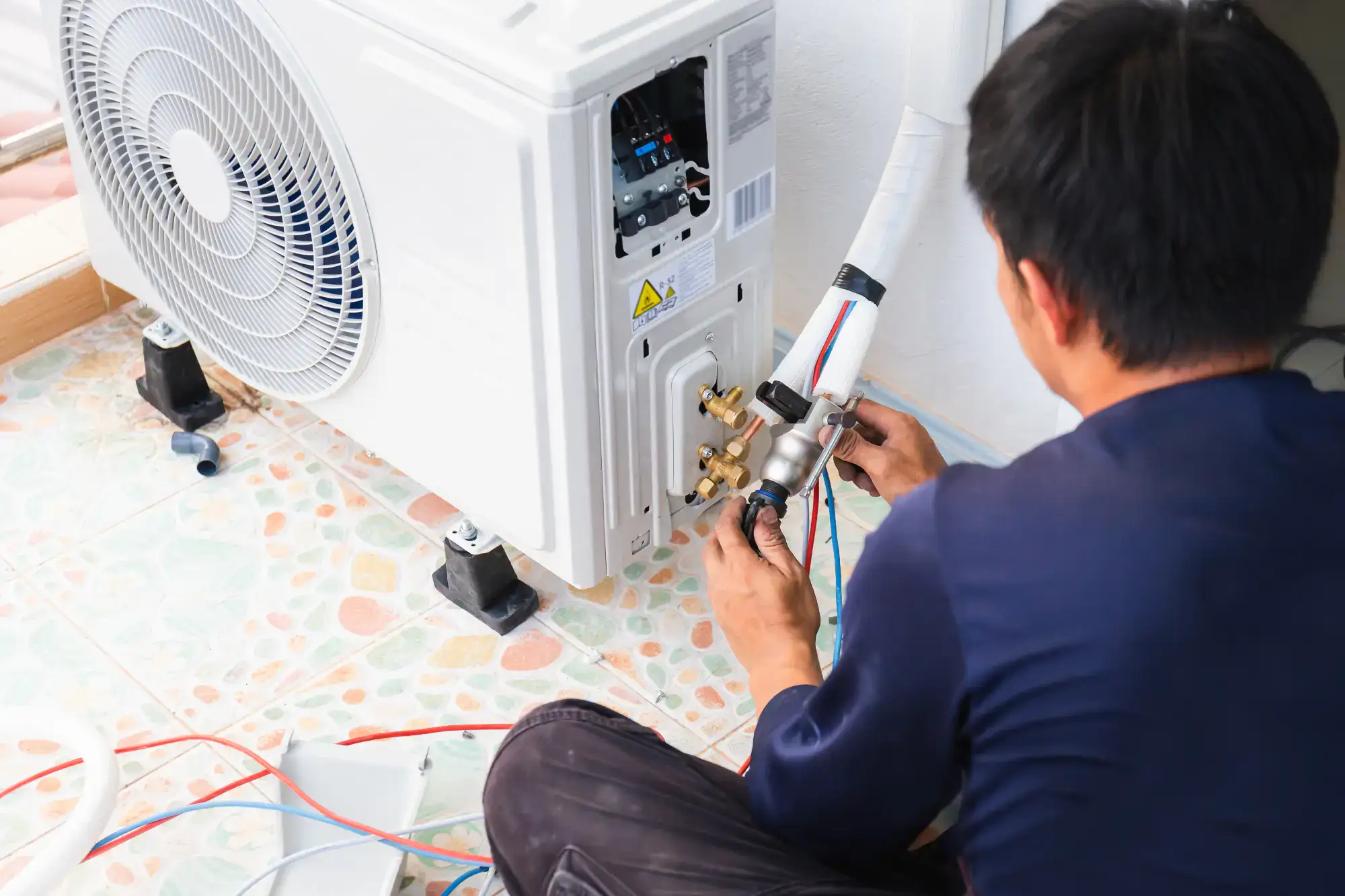 A technician is installing or repairing an air conditioning unit, connecting pipes and wires to the outdoor condenser on a tiled floor.