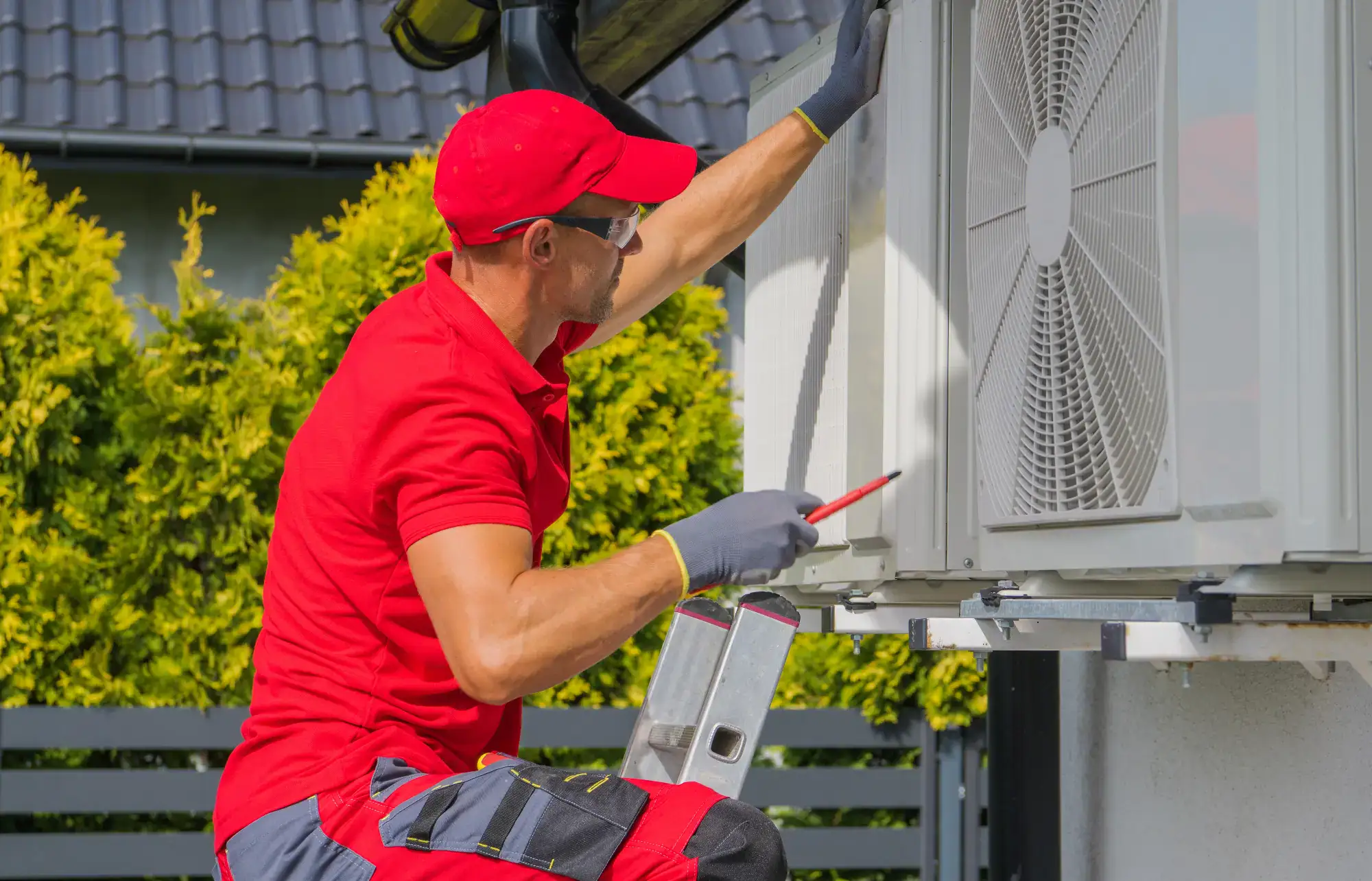 A technician in a red uniform and cap works on an outdoor air conditioning unit with a screwdriver, standing on a small ladder beside a house, with green bushes in the background.