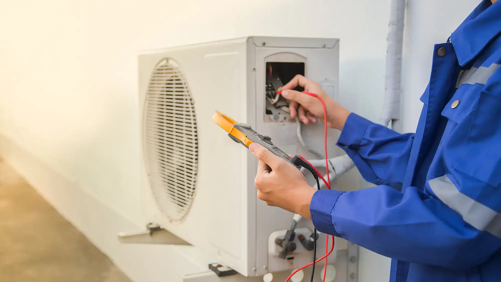 A man wearing blue overalls is skillfully working on an air conditioner in Bergen County, NJ. The technician is focused, crouched next to the unit, and wearing safety gloves as he inspects and repairs the equipment. The background features a residential home, with lush greenery typical of the Bergen County area, showcasing a typical suburban neighborhood.