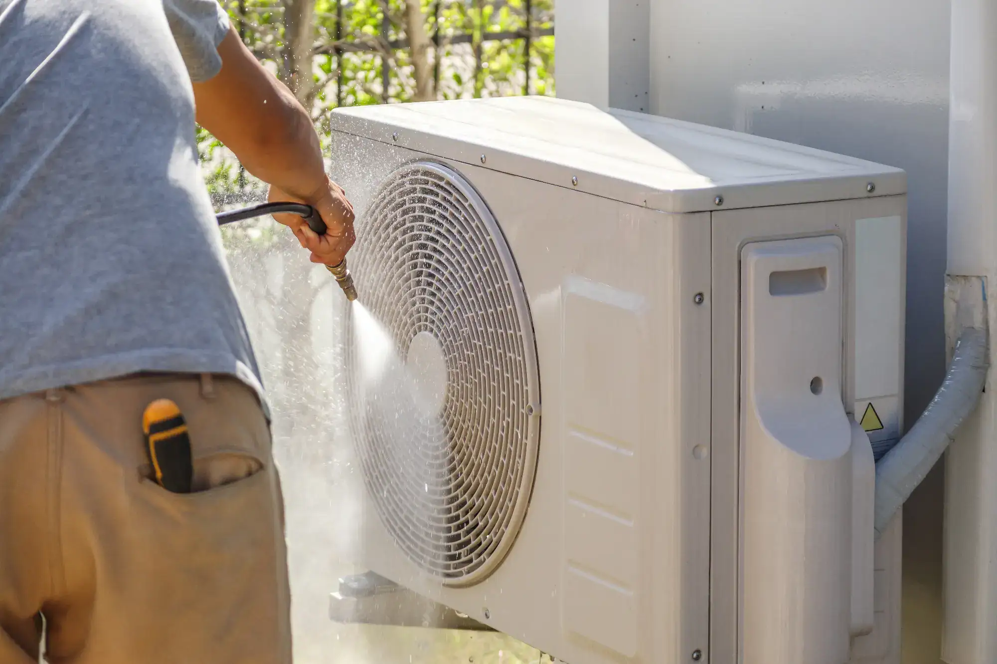 A person uses a pressure washer to clean the outdoor unit of an air conditioner. The unit is mounted outside, and the person is seen from the side wearing a gray shirt and brown pants with tools in the pocket.
