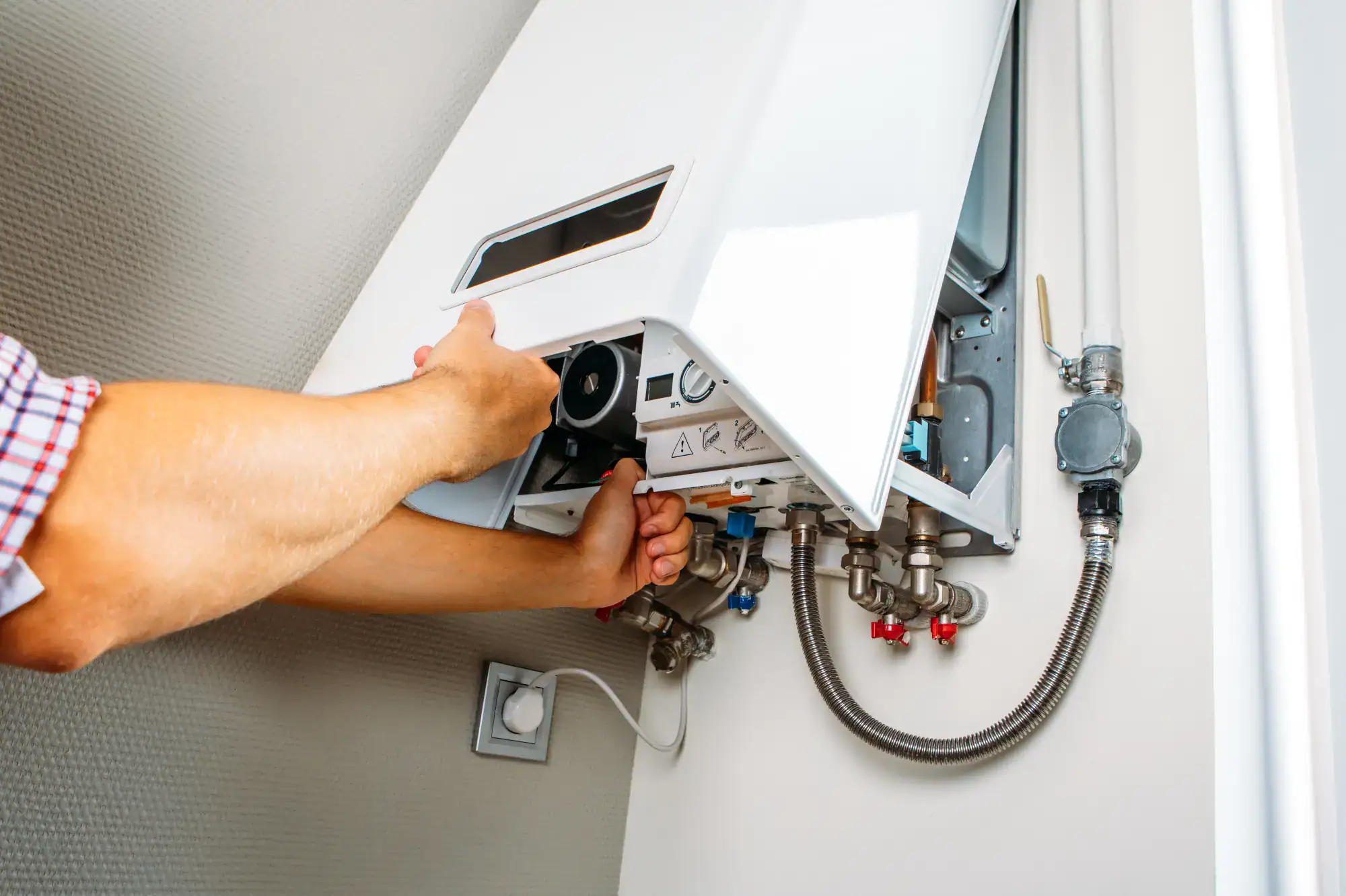 A person inspects or repairs a wall-mounted gas boiler, with the front cover partially open, revealing internal components and various pipes connected below.