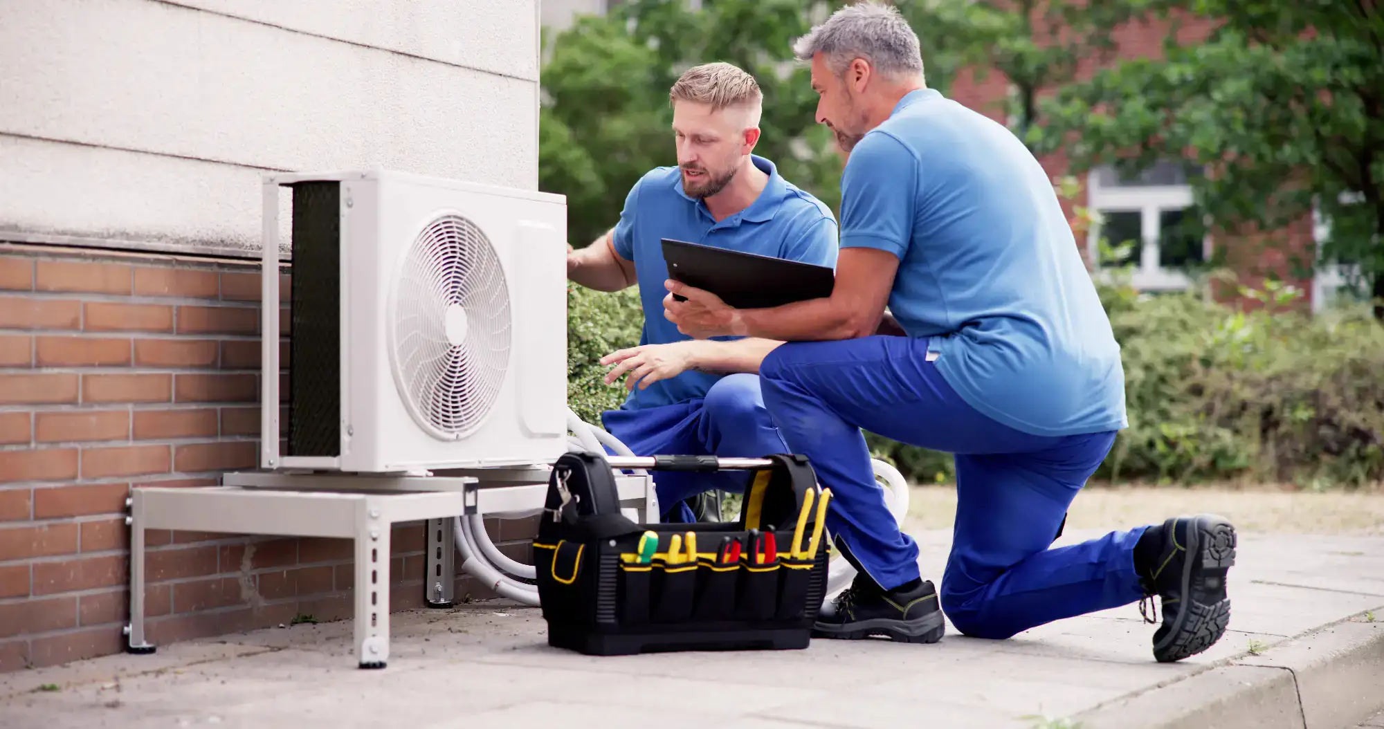 Two technicians in blue uniforms kneel beside an outdoor air conditioning unit, inspecting it with tools and a clipboard, while a black tool bag with various tools sits on the ground nearby.