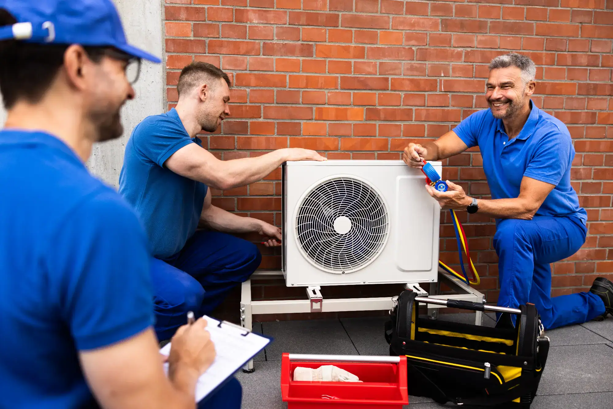 Three professional technicians working together to repair an air conditioner in Bergen County, NJ. They are focused on the unit, with one examining the components, while the others assist with tools and troubleshooting. The work is being done in a residential home with modern furnishings and large windows that let in natural light.