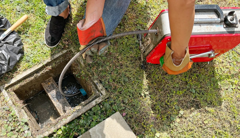 A person wearing gloves uses a plumbing snake machine to clear a clogged outdoor drain, standing on grass next to an open drain cover.