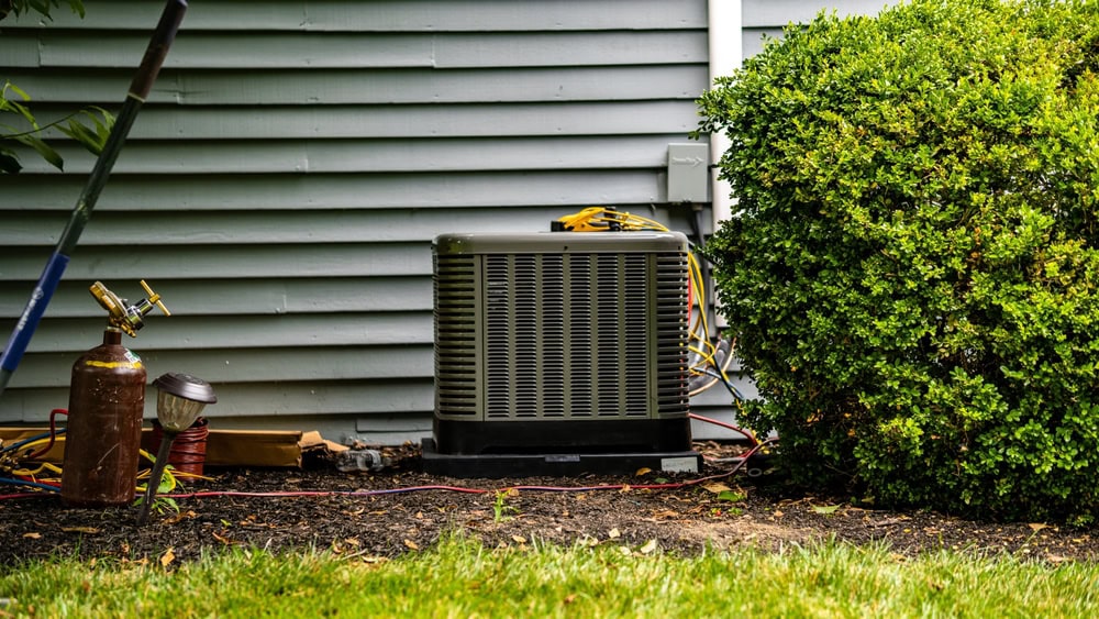 An outdoor air conditioning unit sits on the side of a house, next to a large green bush and a gas tank