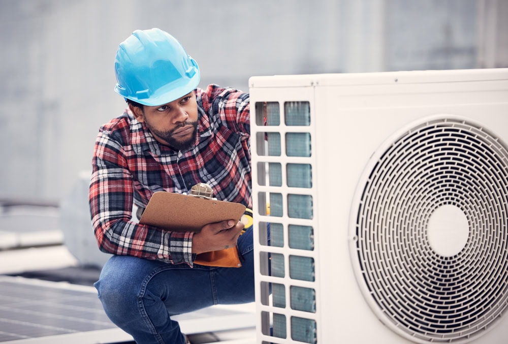 An HVAC technician in a hard hat inspects an outdoor air conditioning unit while holding a clipboard.