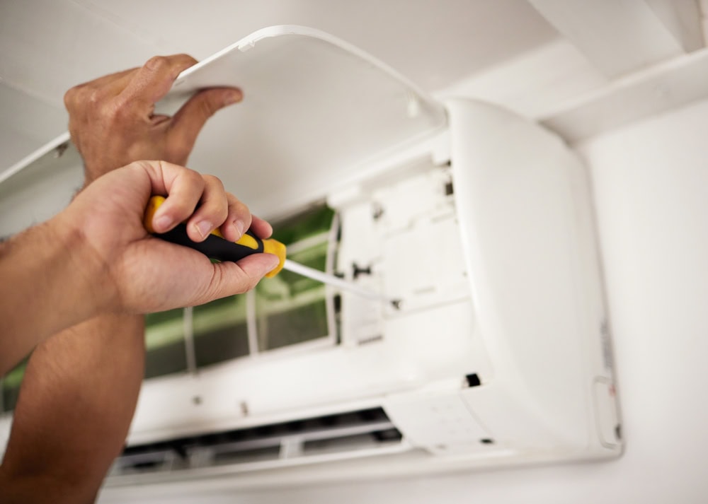 A person uses a screwdriver to repair or maintain a wall-mounted air conditioning unit with its cover open.