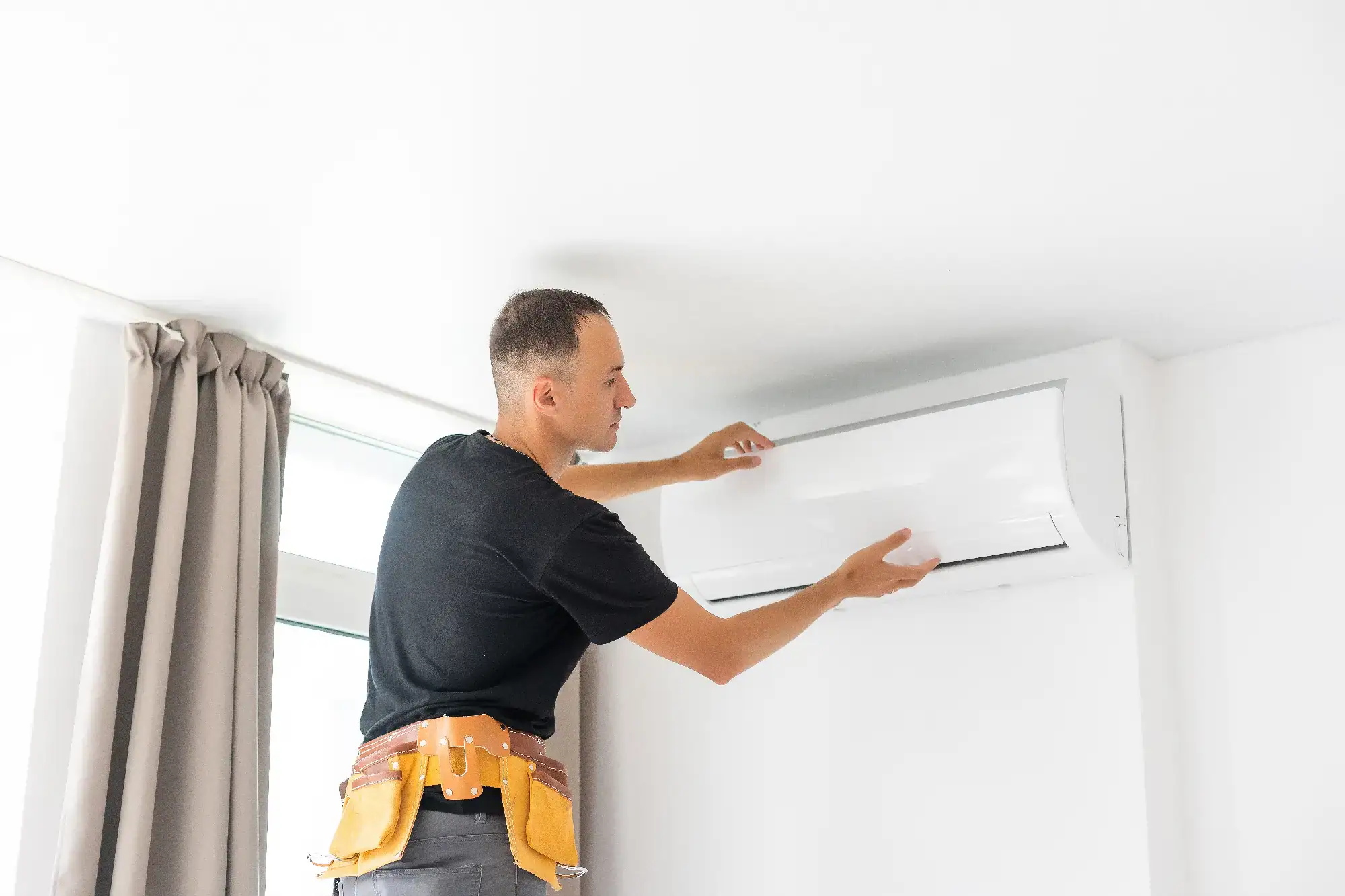 A technician wearing a tool belt adjusts or inspects a wall-mounted air conditioner unit inside a room with white walls and a window with beige curtains.