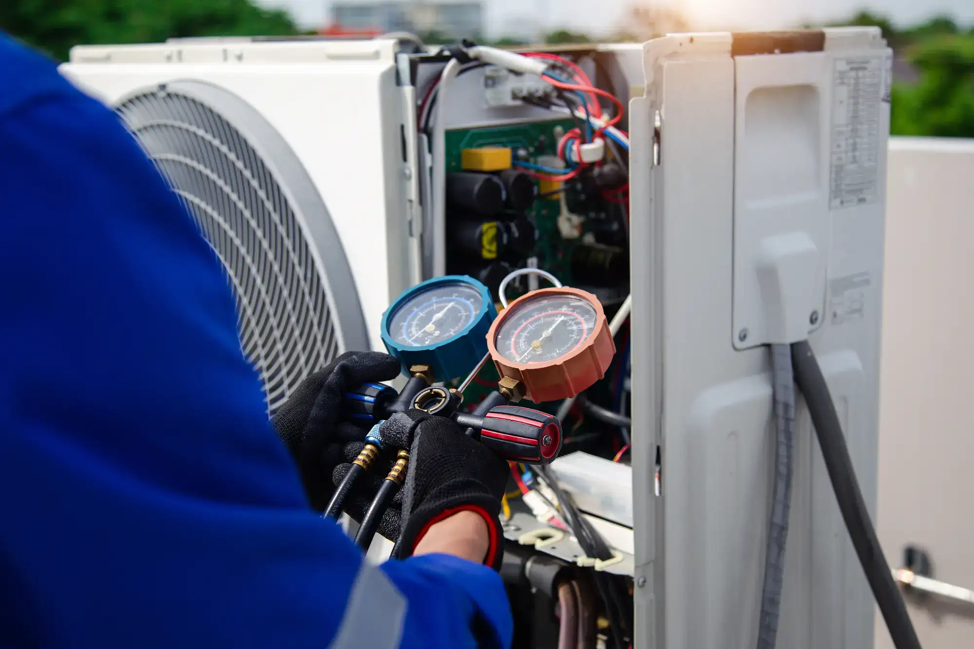 A technician wearing gloves and a blue uniform uses a pressure gauge to check or repair an outdoor air conditioning unit, with wires and components exposed.